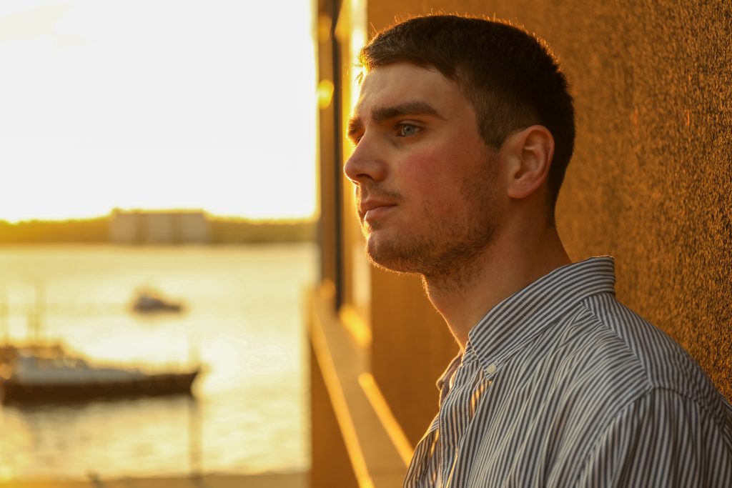 Young man stands leaning against a wall in sunset.